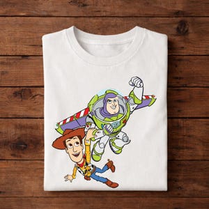 May include: White t-shirt featuring a colorful graphic of Buzz Lightyear and Woody from Toy Story. Buzz Lightyear is in a green and white spacesuit with wings, and Woody is wearing a cowboy hat and vest. The design is centered on the shirt.