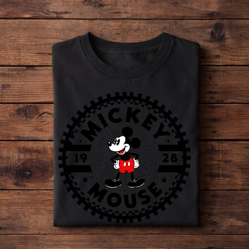 K&ouml;nnte beinhalten: Schwarzes T-Shirt mit einem kreisf&ouml;rmigen Design mit "MICKEY MOUSE" und den Zahlen "1928". Das Design zeigt Mickey Mouse in roten Shorts, schwarzen Schuhen und wei&szlig;en Handschuhen. Das T-Shirt ist auf einer Holzoberfl&auml;che gefaltet.