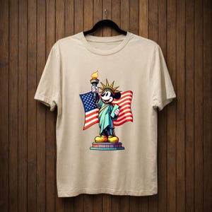 May include: Beige t-shirt featuring a cartoon depiction of Mickey Mouse as the Statue of Liberty, holding a torch and a tablet, with an American flag in the background. The design is colorful and patriotic.