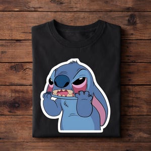 May include: Black t-shirt featuring a cartoon image of Stitch from Lilo & Stitch. The blue alien character has an angry expression, with red-rimmed eyes and a wide open mouth. The shirt is folded on a wooden surface.