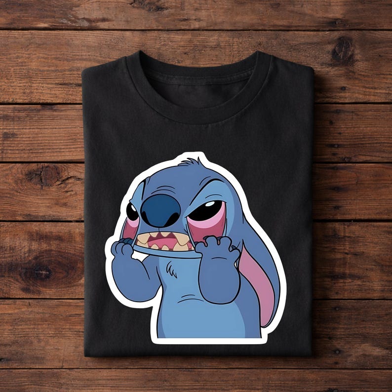 May include: Black t-shirt featuring a cartoon image of Stitch from Lilo & Stitch. The blue alien character has an angry expression, with red-rimmed eyes and a wide open mouth. The shirt is folded on a wooden surface.