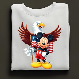May include: White sweatshirt featuring a graphic of Mickey Mouse holding an American flag, with a bald eagle behind him. Mickey is wearing red shorts, yellow shoes, and white gloves. The eagle has brown wings and a white head.