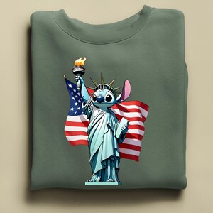 Puede incluir: Una sudadera verde salvia con un personaje de dibujos animados como la Estatua de la Libertad, sosteniendo una antorcha y una bandera estadounidense. El personaje tiene piel azul, ojos grandes y una oreja rosa. La bandera tiene rayas rojas y blancas y estrellas azules.
