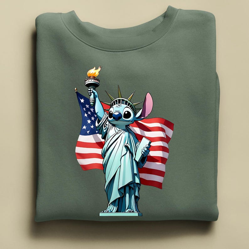 Puede incluir: Una sudadera verde salvia con un personaje de dibujos animados como la Estatua de la Libertad, sosteniendo una antorcha y una bandera estadounidense. El personaje tiene piel azul, ojos grandes y una oreja rosa. La bandera tiene rayas rojas y blancas y estrellas azules.