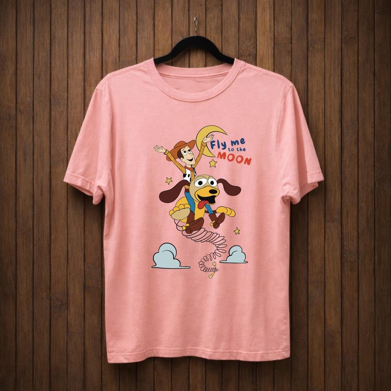 Peut inclure: T-shirt rose clair avec une illustration de dessin anim&eacute; d'un cow-boy et d'un chien, avec le texte "Fly me to the moon". Le design montre les personnages volant vers une lune en croissant. Le t-shirt est suspendu &agrave; un cintre noir sur un fond en bois.