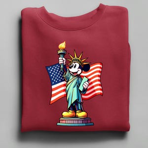 Puede incluir: Una sudadera granate con una ilustraci&oacute;n de dibujos animados de Mickey Mouse como la Estatua de la Libertad, sosteniendo una antorcha y una tableta, con una bandera estadounidense de fondo. El dise&ntilde;o es colorido y patri&oacute;tico.