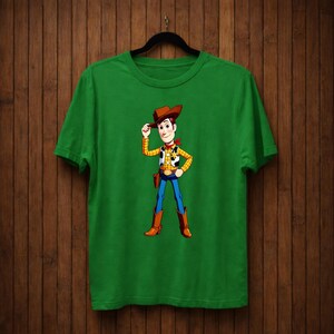 May include: A green t-shirt with a cartoon cowboy character illustration. The character wears a brown cowboy hat, yellow vest, red neckerchief, blue jeans, and brown boots. The t-shirt is displayed on a black hanger.