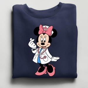 Puede incluir: Sudadera azul marino con Minnie Mouse vestida de enfermera. Minnie Mouse lleva una bata blanca, un lazo rosa y un estetoscopio. Est&aacute; haciendo un gesto de coraz&oacute;n con los dedos. La sudadera es de color azul marino liso.