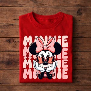 K&ouml;nnte beinhalten: Ein rotes T-Shirt mit einer Minnie Mouse-Grafik, die eine herzf&ouml;rmige Sonnenbrille und eine Schleife tr&auml;gt. Das Wort "MINNIE" ist vertikal in Rosa und Wei&szlig; gedruckt. Das T-Shirt ist auf einer Holzoberfl&auml;che gefaltet.