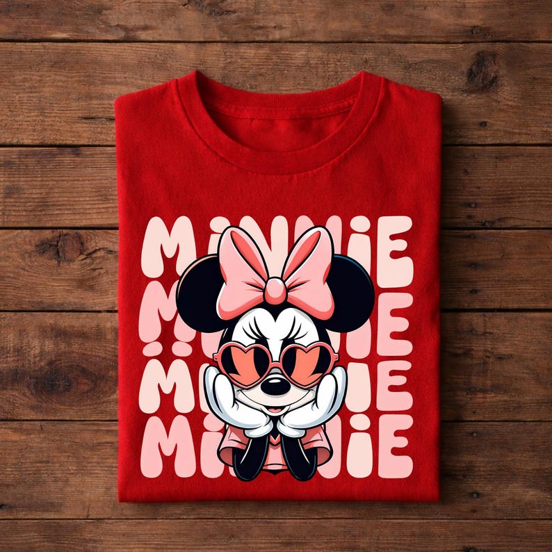 K&ouml;nnte beinhalten: Ein rotes T-Shirt mit einer Minnie Mouse-Grafik, die eine herzf&ouml;rmige Sonnenbrille und eine Schleife tr&auml;gt. Das Wort "MINNIE" ist vertikal in Rosa und Wei&szlig; gedruckt. Das T-Shirt ist auf einer Holzoberfl&auml;che gefaltet.