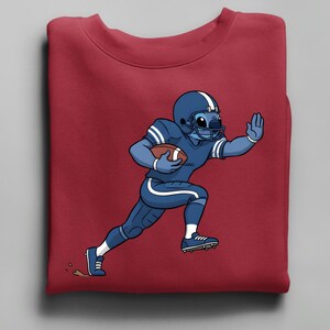 May include: A maroon sweatshirt with a cartoon character in a blue American football uniform, including a helmet and pads, running with a football. The character is light blue with white stripes.