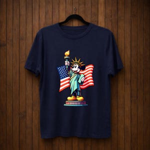 May include: Navy blue t-shirt featuring a cartoon character dressed as the Statue of Liberty, holding a torch and a tablet, with an American flag in the background. The design is colorful and patriotic.