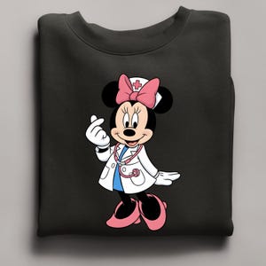 Puede incluir: Sudadera negra con Minnie Mouse vestida de enfermera. Minnie lleva una bata blanca, un lazo rosa y un estetoscopio. Est&aacute; haciendo un coraz&oacute;n con los dedos. El dise&ntilde;o est&aacute; centrado en la sudadera.