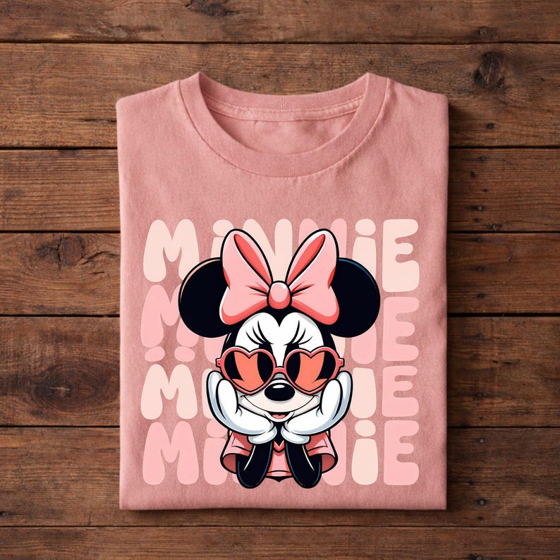 K&ouml;nnte beinhalten: Ein gefaltetes rosa T-Shirt mit einem Minnie Mouse-Motiv. Minnie tr&auml;gt herzf&ouml;rmige Sonnenbrillen und eine rosa Schleife. Das Wort "MINNIE" ist in einem sich wiederholenden Muster in einer helleren rosa Farbe gedruckt.