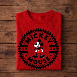 K&ouml;nnte beinhalten: Ein gefaltetes rotes T-Shirt mit einer schwarz-wei&szlig;en Mickey Mouse-Grafik. Das Design enth&auml;lt die W&ouml;rter "MICKEY MOUSE" und die Zahlen "19" und "28" in einem kreisf&ouml;rmigen Design.