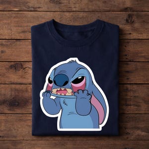 May include: Navy blue t-shirt featuring a cartoon image of Stitch from Lilo & Stitch. The character is blue with large ears, a wide mouth, and pink accents. The image is printed on a folded t-shirt.