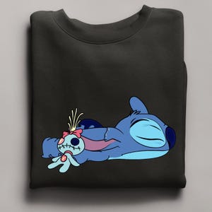 Puede incluir: Sudadera negra con una ilustraci&oacute;n de dibujos animados de Stitch, un personaje alien&iacute;gena azul, acostado y sosteniendo una mu&ntilde;eca. El gr&aacute;fico es en tonos de azul, rosa y blanco. La sudadera es de color negro liso.