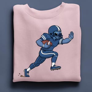 May include: A pale pink sweatshirt with a cartoon character in a blue American football uniform, running with a brown football. The character is wearing a helmet and has a hand raised. The design is centered on the sweatshirt.