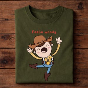 Puede incluir: Camiseta verde oliva con una ilustraci&oacute;n de dibujos animados de un personaje de vaquero con sombrero marr&oacute;n, camisa amarilla, vaqueros azules y botas marrones. El texto "Feelin woody" est&aacute; escrito en rojo encima del personaje.