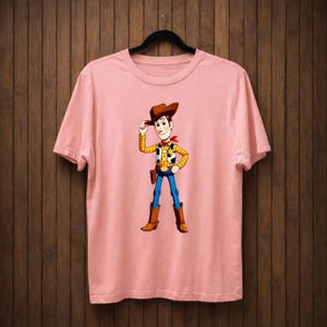 May include: A pale pink t-shirt with a cartoon cowboy character. The cowboy wears a brown hat, yellow plaid vest, blue jeans, and brown boots. A red bandana is around his neck. The t-shirt is hanging on a black hanger.