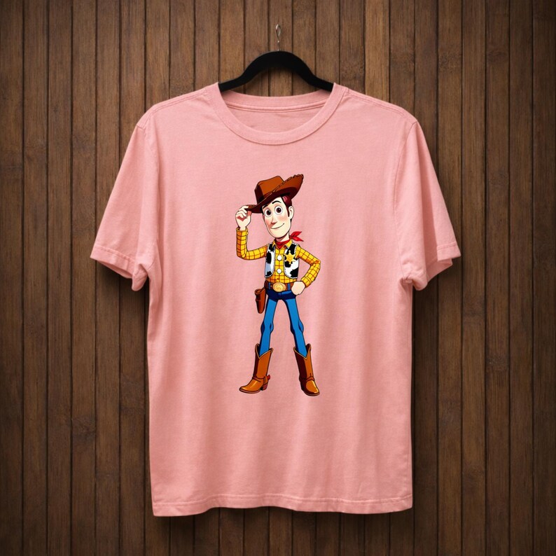 May include: A pale pink t-shirt with a cartoon cowboy character. The cowboy wears a brown hat, yellow plaid vest, blue jeans, and brown boots. A red bandana is around his neck. The t-shirt is hanging on a black hanger.