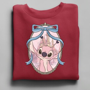 Puede incluir: Una sudadera granate con un personaje de dibujos animados rosa dentro de un marco ovalado. El marco est&aacute; adornado con un lazo azul claro y detalles de coraz&oacute;n. El fondo muestra un castillo. La sudadera est&aacute; doblada.