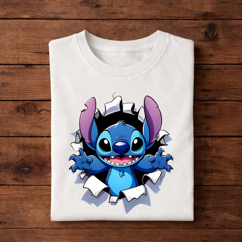 May include: White t-shirt featuring a cartoon illustration of Stitch, a blue alien character, bursting through a torn hole. Stitch has large ears, a wide smile, and outstretched arms. The design is centered on the shirt.