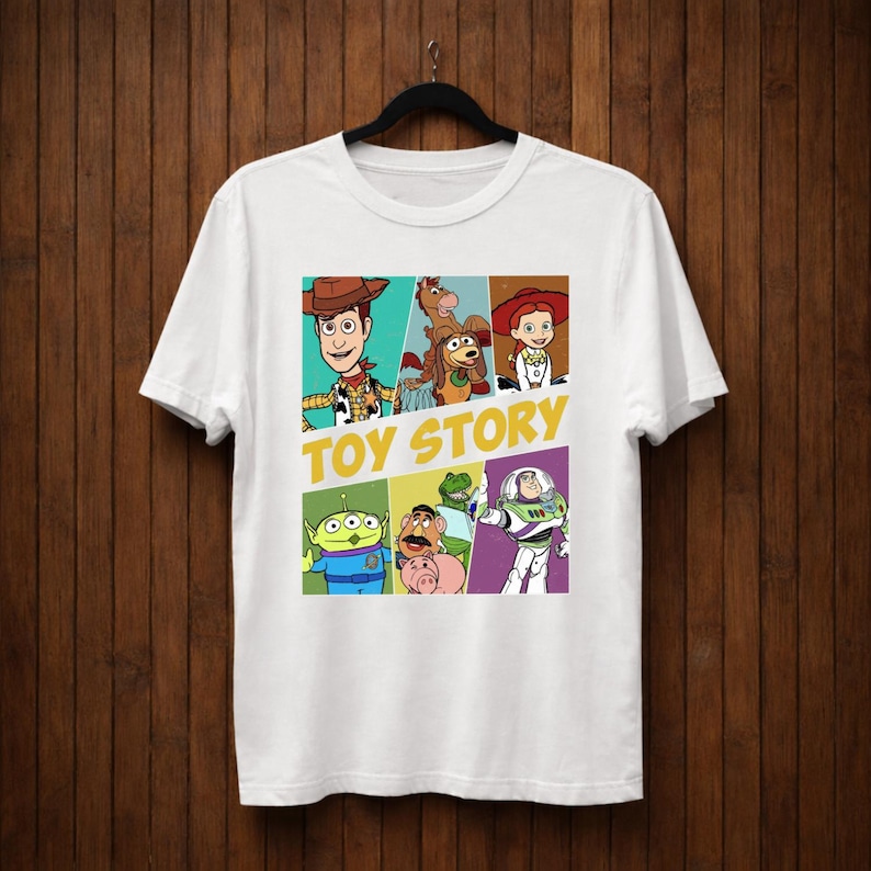 K&ouml;nnte beinhalten: Wei&szlig;es T-Shirt mit einer farbenfrohen Grafik von Charakteren aus dem Animationsfilm "Toy Story". Das Design umfasst Woody, Buzz Lightyear und andere Charaktere, mit den Worten "Toy Story" in Gelb.