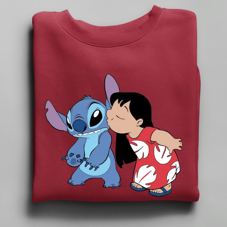 Peut inclure: Un sweat-shirt bordeaux avec une illustration de Stitch et Lilo. Stitch est bleu, et Lilo porte une robe rouge &agrave; motifs de feuilles blanches. Lilo embrasse Stitch sur la joue. Le sweat-shirt est pli&eacute;.