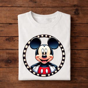 May include: White t-shirt featuring a colorful graphic of Mickey Mouse. The cartoon character is inside a black and white striped circle. Mickey Mouse is wearing a red and white outfit with black gloves and shoes. The shirt is folded on a wooden surface.