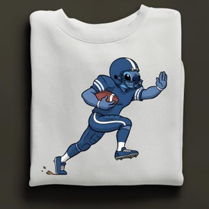 May include: White sweatshirt with a cartoon character in a blue American football uniform, holding a brown ball. The character is running, wearing a blue helmet and shoes. A fun design for sports fans.