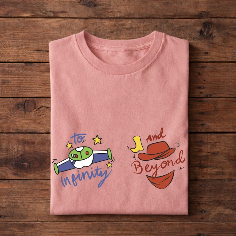Op de afbeelding: Een gevouwen, lichtroze t-shirt met de tekst "To Infinity and Beyond" in een speels lettertype. Het ontwerp bevat een cartoon ruimteschip, een cowboyhoed en een laars.