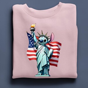 Puede incluir: Sudadera rosa claro con un personaje de dibujos animados como la Estatua de la Libertad, sosteniendo una antorcha y una tableta, con una bandera estadounidense de fondo. El personaje es azul con ojos y orejas grandes.