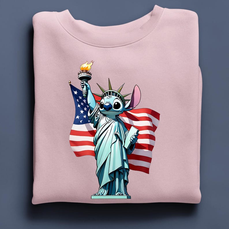 Puede incluir: Sudadera rosa claro con un personaje de dibujos animados como la Estatua de la Libertad, sosteniendo una antorcha y una tableta, con una bandera estadounidense de fondo. El personaje es azul con ojos y orejas grandes.