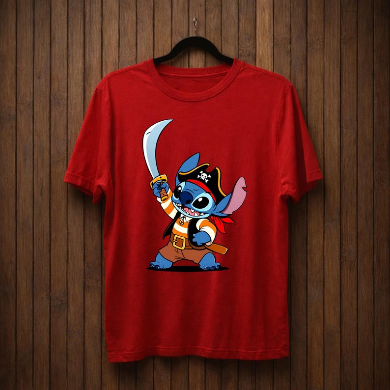May include: Red t-shirt featuring a cartoon character dressed as a pirate, holding a sword. The character is blue with large eyes, wearing a pirate hat, vest, and red bandana. The shirt is hanging on a black hanger against a wooden background.