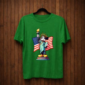 May include: Green t-shirt featuring a cartoon character dressed as the Statue of Liberty, holding a torch and a tablet, with an American flag in the background. The design is centered on the shirt.