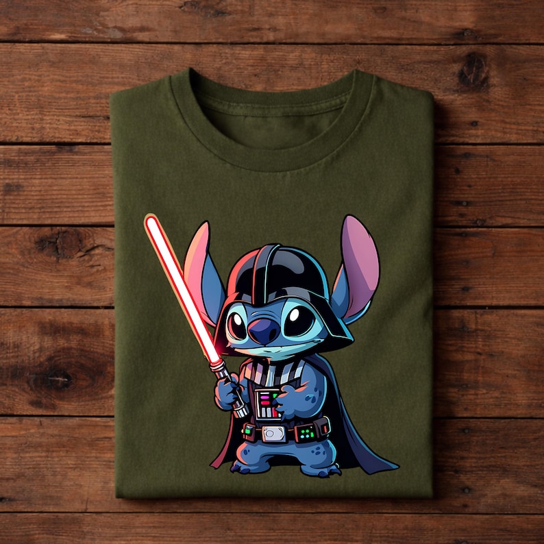 Pu&ograve; includere: T-shirt verde oliva con un personaggio dei cartoni animati vestito da Darth Vader, con una spada laser rossa. Il personaggio &egrave; blu con grandi occhi, indossa un casco e un mantello neri. Il disegno &egrave; centrato sulla maglietta piegata.