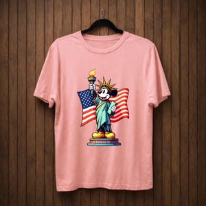 May include: A light pink t-shirt featuring a cartoon depiction of Mickey Mouse as the Statue of Liberty, holding a torch and a tablet, with an American flag in the background. The design is colorful and detailed.