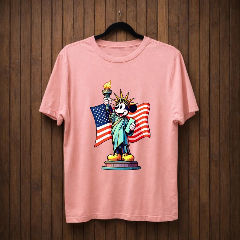 May include: A light pink t-shirt featuring a cartoon depiction of Mickey Mouse as the Statue of Liberty, holding a torch and a tablet, with an American flag in the background. The design is colorful and detailed.