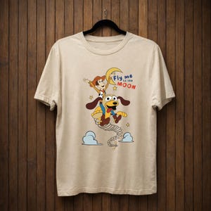 Peut inclure: T-shirt beige avec un dessin anim&eacute; de Woody et d'un chien volant vers la lune. L'inscription "Fly me to the Moon" est au-dessus des personnages en bleu et rouge. Le motif comprend des &eacute;toiles et des nuages.