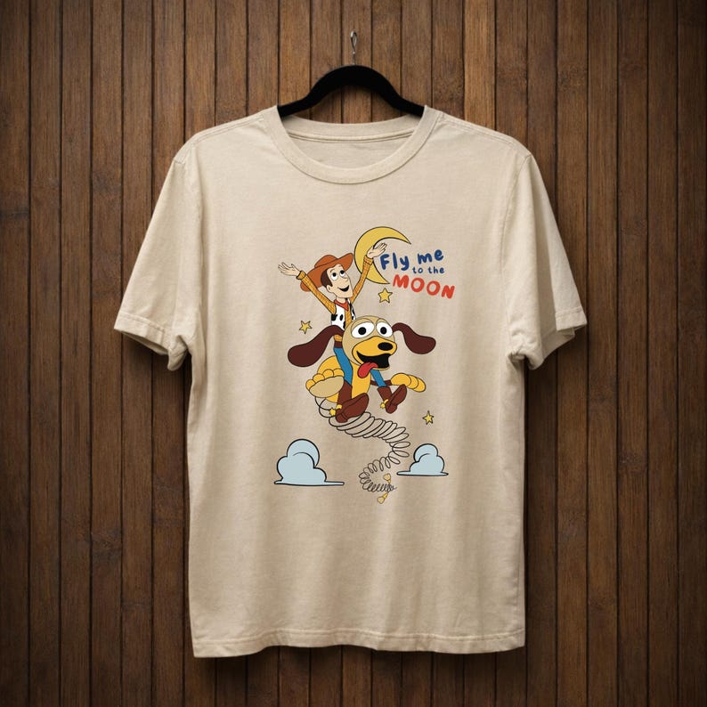 Peut inclure: T-shirt beige avec un dessin anim&eacute; de Woody et d'un chien volant vers la lune. L'inscription "Fly me to the Moon" est au-dessus des personnages en bleu et rouge. Le motif comprend des &eacute;toiles et des nuages.