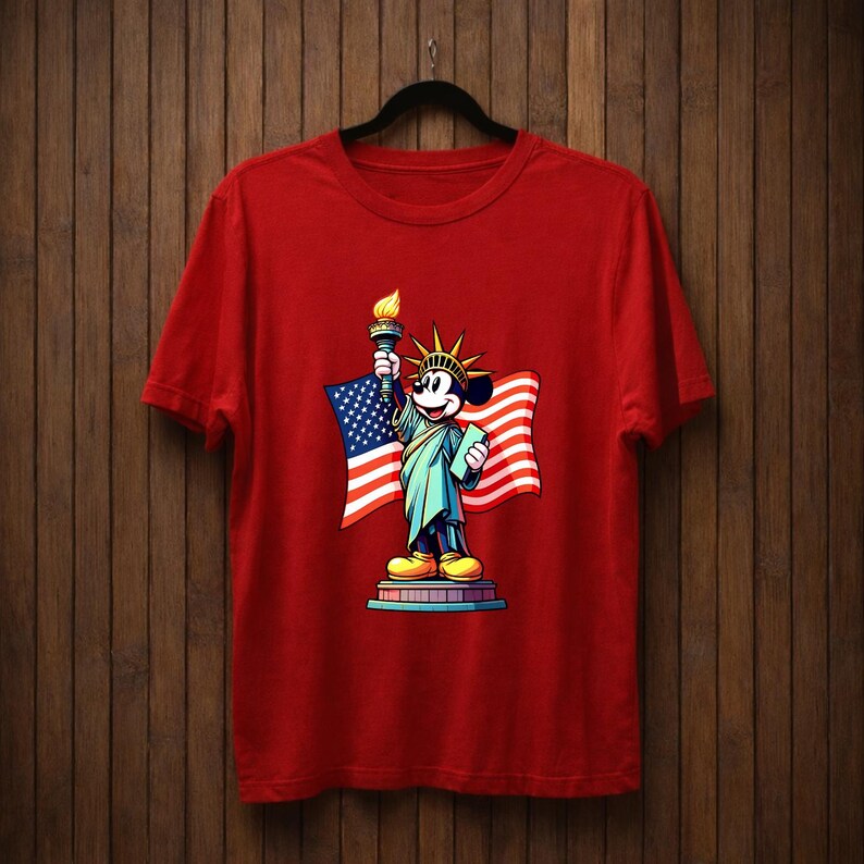May include: Red t-shirt featuring a graphic of Mickey Mouse dressed as the Statue of Liberty, holding a torch and a tablet, with an American flag in the background. The design is colorful and patriotic.