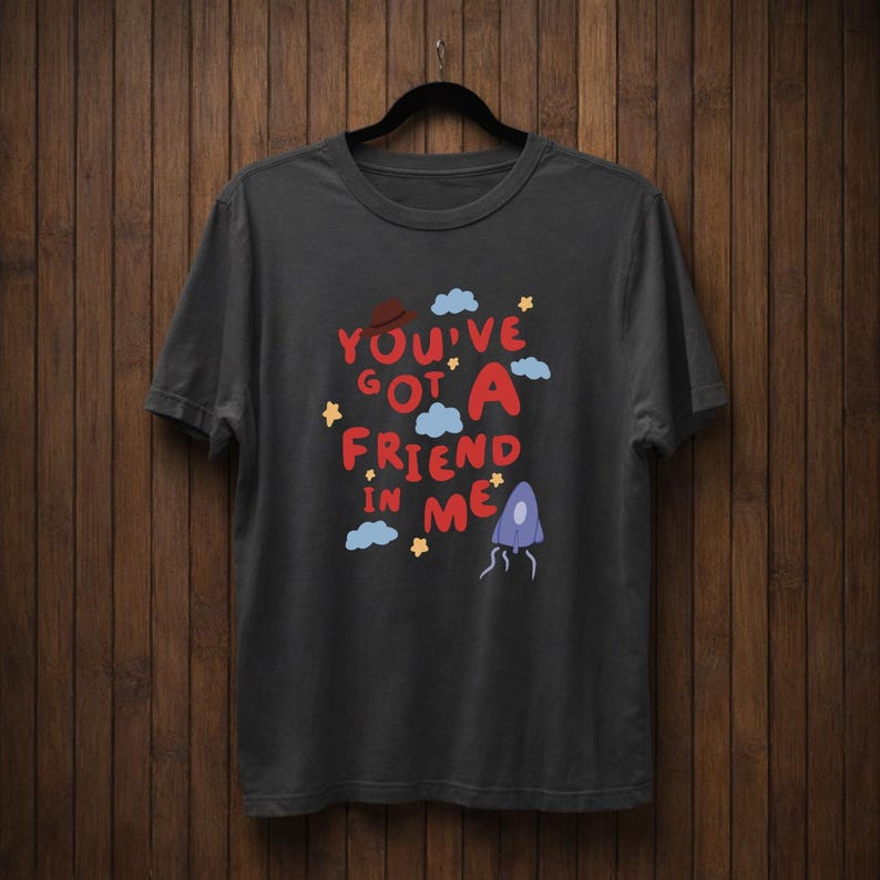 Op de afbeelding: Donkergrijs t-shirt met de tekst "You've Got A Friend In Me" in rood, bruin en blauw. Het ontwerp bevat sterren, wolken en een grillig karakter. Het t-shirt hangt aan een zwarte hanger.