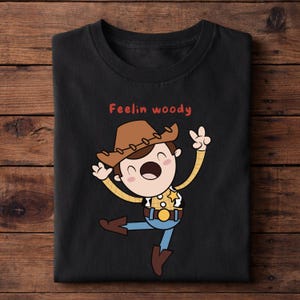Puede incluir: Camiseta negra con una ilustraci&oacute;n de dibujos animados de un vaquero con sombrero marr&oacute;n, camisa amarilla, pantalones azules y botas marrones. El texto "Feelin woody" en rojo est&aacute; encima.