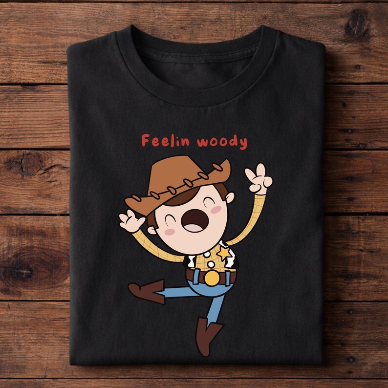 Puede incluir: Camiseta negra con una ilustraci&oacute;n de dibujos animados de un vaquero con sombrero marr&oacute;n, camisa amarilla, pantalones azules y botas marrones. El texto "Feelin woody" en rojo est&aacute; encima.