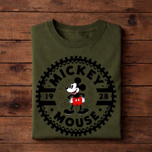 K&ouml;nnte beinhalten: Olivgr&uuml;nes T-Shirt mit einem schwarzen Mickey Mouse-Motiv und dem Text "MICKEY MOUSE 1928". Das Design ist kreisf&ouml;rmig mit einem zahnradartigen Rand. Das T-Shirt ist auf einer Holzoberfl&auml;che gefaltet.