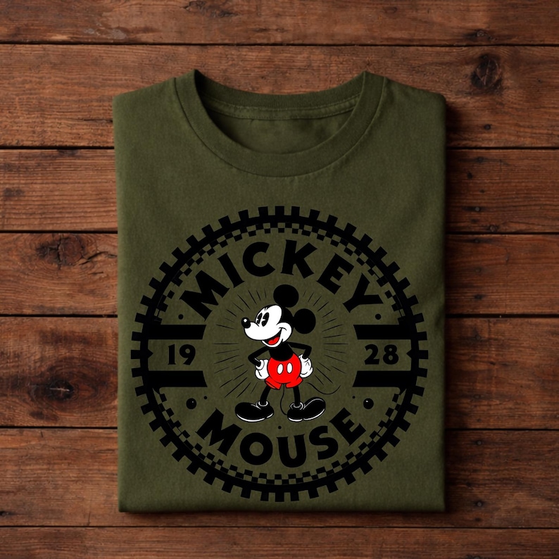 K&ouml;nnte beinhalten: Olivgr&uuml;nes T-Shirt mit einem schwarzen Mickey Mouse-Motiv und dem Text "MICKEY MOUSE 1928". Das Design ist kreisf&ouml;rmig mit einem zahnradartigen Rand. Das T-Shirt ist auf einer Holzoberfl&auml;che gefaltet.