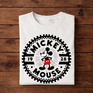 K&ouml;nnte beinhalten: Wei&szlig;es T-Shirt mit einem kreisf&ouml;rmigen Mickey Mouse-Motiv. Das Design enth&auml;lt die W&ouml;rter "MICKEY MOUSE" und die Zahlen "19" und "28". Mickey Mouse ist in seiner klassischen Pose zu sehen, mit roter Hose und schwarzen Schuhen.