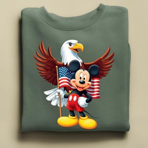May include: A sage green sweatshirt featuring a graphic of Mickey Mouse holding an American flag, with a bald eagle behind him. Mickey is wearing red shorts, black gloves, and yellow shoes. The eagle has brown wings and a white head.