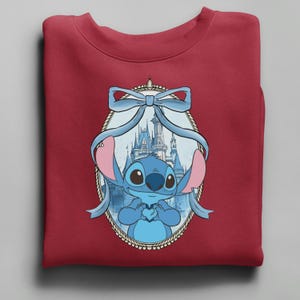 K&ouml;nnte beinhalten: Ein weinrotes Sweatshirt mit einer blauen Cartoonfigur, die ein Herz h&auml;lt, umrahmt von einer hellblauen Schleife und einem Schloss im Hintergrund. Die Figur hat gro&szlig;e Augen und rosa Ohren. Das Design ist auf dem gefalteten Kleidungsst&uuml;ck zentriert.